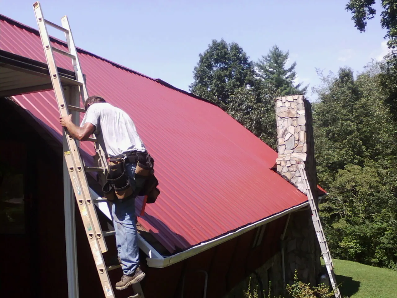 Roofer on a ladder beside a red metal roof and stone chimney in Norborne